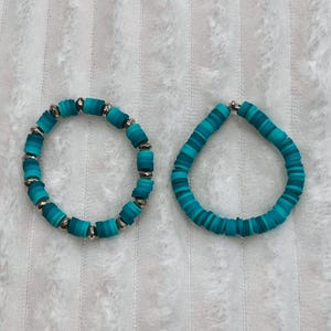 May include: Two turquoise and teal beaded bracelets. One bracelet features cylindrical beads with silver accents, while the other has flat, disc-shaped beads. The bracelets are displayed on a white, textured surface.