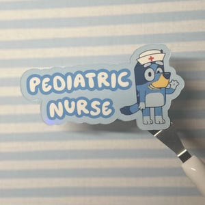 May include: A light blue sticker with the words "PEDIATRIC NURSE" in white. The sticker features a cartoon dog wearing a nurse's hat. The dog is blue with a yellow nose and is waving.