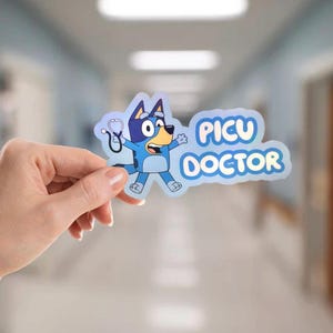 Puede incluir: Una pegatina con un personaje de perro azul de dibujos animados sosteniendo un estetoscopio. La pegatina tiene las palabras "PICU DOCTOR" en texto azul. El fondo es un pasillo de hospital borroso.