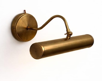 May include: A brass-toned wall-mounted picture light. The light features a circular base, a curved arm, and a cylindrical light fixture. The fixture is designed to illuminate artwork or photographs. The finish is a warm, metallic tone.