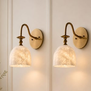 May include: A pair of wall sconces featuring circular brass backplates, curved brass arms, and dome-shaped white alabaster shades. Each fixture emits a warm, downward-facing light, showcasing the natural veining and translucent texture of the stone.