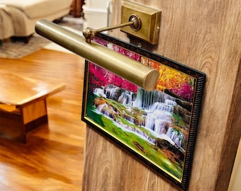 May include: A brass picture light illuminates a framed landscape print of a waterfall. The light is mounted on a wooden surface. The print features vibrant colors of green, pink, and yellow.