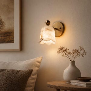 May include: A wall-mounted light fixture with a floral-shaped shade. The shade is white with brown marbling, and the fixture has a brass-colored base and stem. The light is on, casting a warm glow. The fixture is mounted on a beige wall.