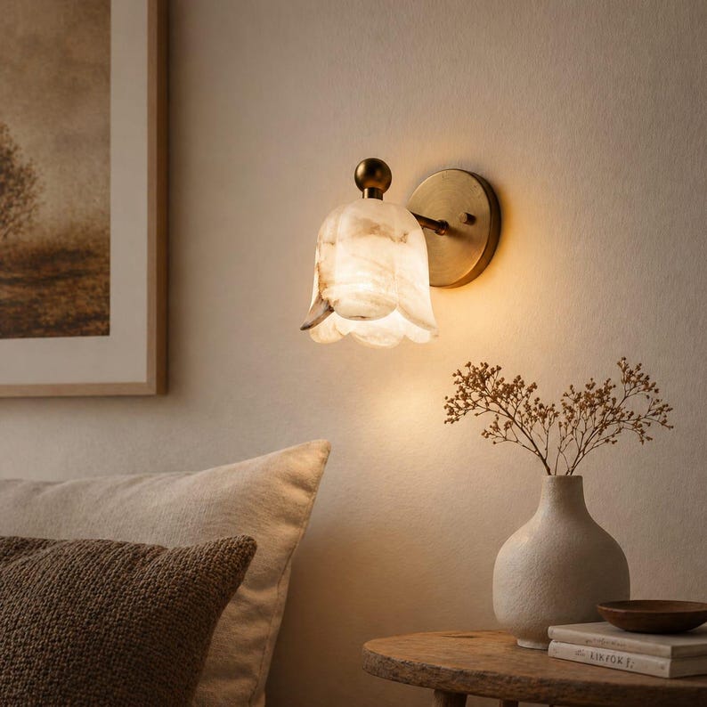 May include: A wall-mounted light fixture with a floral-shaped shade. The shade is white with brown marbling, and the fixture has a brass-colored base and stem. The light is on, casting a warm glow. The fixture is mounted on a beige wall.