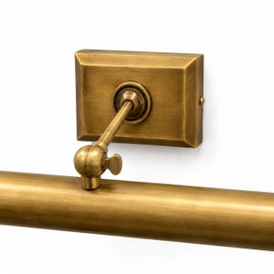 May include: A wall-mounted picture light fixture with a brushed brass finish. The design features a rectangular backplate, a single adjustable arm with a swivel joint, and a long cylindrical light housing with a side-mounted adjustment knob.