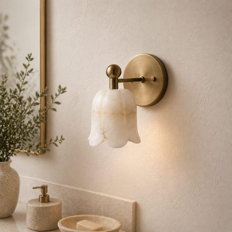May include: A wall-mounted light fixture with a tulip-shaped, off-white shade. The fixture has a brass-colored base and arm. The light is on, casting a warm glow. The fixture is mounted on a light-colored wall, suggesting a bathroom or vanity setting.