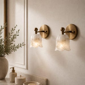 May include: Two wall-mounted sconces with a tulip-shaped, off-white shade and a brass-colored base. The sconces are mounted on a light-colored wall. The shades are translucent, allowing light to shine through. Decorative lighting.