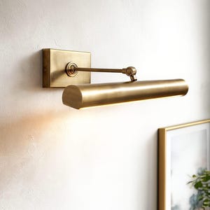 May include: A brass-toned wall-mounted picture light. The fixture has a rectangular backplate, an adjustable arm, and a cylindrical light housing. The light is designed to illuminate artwork and has a warm glow.
