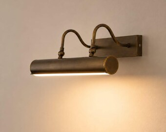 May include: A wall-mounted picture light with an antique brass finish. The fixture features a rectangular backplate, two curved arms, and a cylindrical lamp head that emits a warm, downward-facing light along its length.