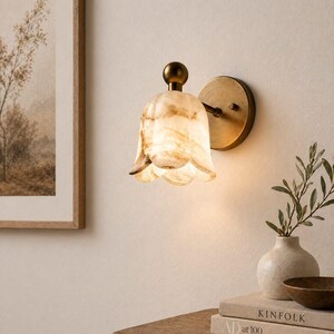 May include: A wall-mounted light fixture with a tulip-shaped shade made of translucent, cream-colored stone with brown veining. The fixture has a brass-colored base and a small, round finial. The light is on, casting a warm glow.