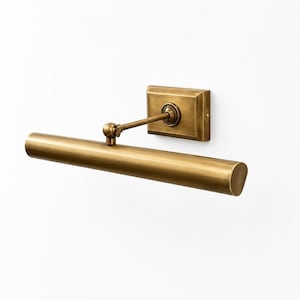 May include: A wall-mounted picture light fixture with a brushed brass finish. The design features a rectangular backplate, a single adjustable arm with a pivot joint, and a long, cylindrical horizontal light housing for illuminating artwork.
