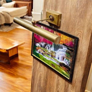 May include: A brass picture light mounted above a framed landscape print featuring a multi-tiered waterfall with vibrant green foliage and pink-toned trees. The rectangular frame has a dark, textured finish and is attached to a wooden surface.