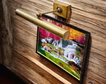 May include: A brass picture light is mounted above a framed landscape painting. The painting depicts a waterfall scene with vibrant colors. The light fixture has a rectangular base and a cylindrical arm, adding a touch of elegance to the artwork.