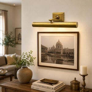 May include: A brass picture light fixture with a cylindrical shade and a rectangular wall mount, positioned above a framed black and white landscape photograph of a domed building near water, mounted on a plain off-white wall surface.