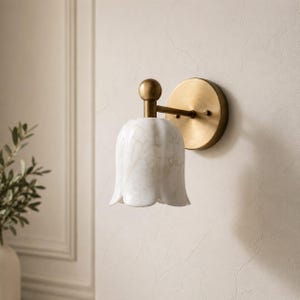 May include: A wall-mounted light fixture with a white, tulip-shaped shade and a brass-colored base. The shade has a marble-like pattern. The fixture is mounted on a beige wall, suggesting a home decor item.