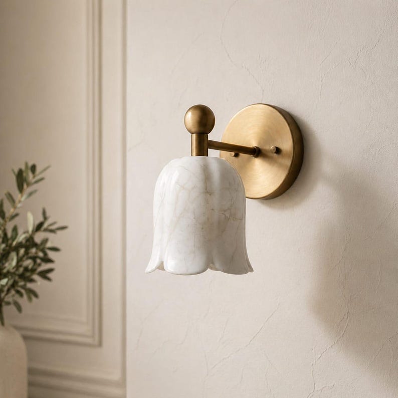 May include: A wall-mounted light fixture with a white, tulip-shaped shade and a brass-colored base. The shade has a marble-like pattern. The fixture is mounted on a beige wall, suggesting a home decor item.