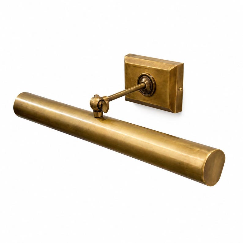 May include: A wall-mounted picture light with a cylindrical brass lamp head and a rectangular mounting plate. The fixture features an adjustable arm with a swivel joint, finished in a brushed antique brass tone with a metallic texture.