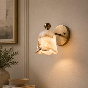 May include: A wall-mounted sconce with a tulip-shaped shade made of translucent, veined stone. The fixture has a brass-colored base and a small, round finial. The shade is a creamy white with brown and tan veining, and the light shines through the stone.
