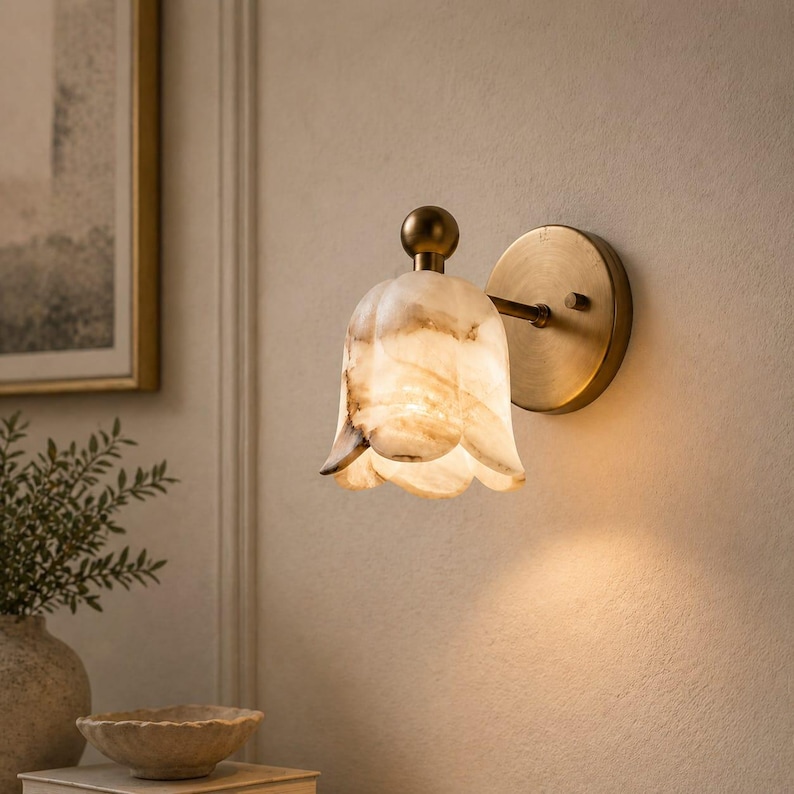 May include: A wall-mounted sconce with a tulip-shaped shade made of translucent, veined stone. The fixture has a brass-colored base and a small, round finial. The shade is a creamy white with brown and tan veining, and the light shines through the stone.