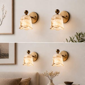 May include: Four wall sconces with a floral design. Each sconce features a cream-colored, petal-shaped shade with a marbled effect, a brass-colored backplate, and a small, round brass finial. The sconces are mounted on a light-colored wall.