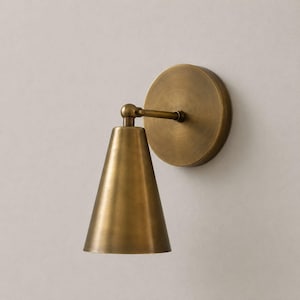 Puede incluir: Un aplique de pared con acabado en latón cepillado. La lámpara cuenta con una placa circular de montaje y una pantalla cónica unida a un brazo corto y ajustable con una articulación giratoria, que dirige la luz hacia abajo.
