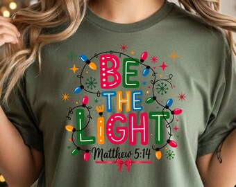 Peut inclure: Un t-shirt vert olive avec le texte "BE THE LIGHT" en lettres colorées et des guirlandes lumineuses de Noël. Le texte "Matthew 5:14" est en dessous. Le t-shirt est de style décontracté et confortable.