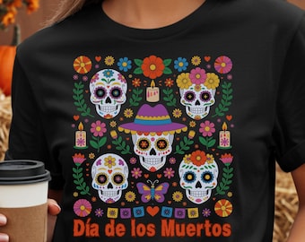 May include: Black t-shirt featuring a colorful Día de los Muertos design with sugar skulls, flowers, and butterflies. The shirt is worn with denim shorts. Pumpkins and hay bales are in the background.