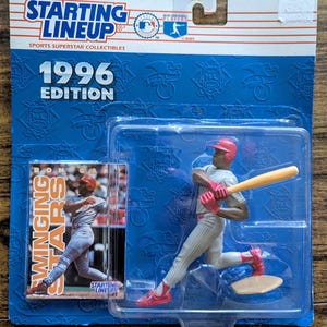 May include: A 1996 Edition Starting Lineup baseball figure of a player in a batting pose, packaged in a blue blister pack. The figure is wearing a red helmet, red shoes, and a gray uniform. A card with the text "Swinging Stars" is included.