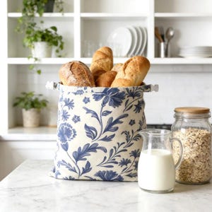 Könnte beinhalten: Ein Stoff-Brotkorb mit blauem Blumenmuster, gefüllt mit verschiedenen Brotsorten. Ein Glaskrug mit Milch und ein Glas mit Haferflocken stehen neben dem Korb. Die Szene spielt in einer Küche mit weißen Regalen und Pflanzen.