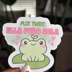 May include: A sticker featuring a cartoon frog on a lily pad with the text "PLOT TWIST: ELLA PUDO SOLA" in green and pink. The frog is light green with pink cheeks and hearts around it.