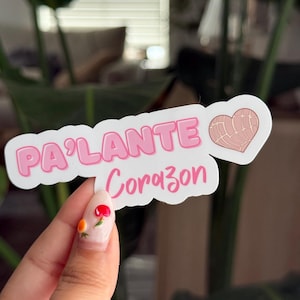 May include: A white sticker with the words "PA'LANTE Corazon" in pink lettering. A heart-shaped concha pastry illustration is on the right. The sticker is held in front of a blurred green background.