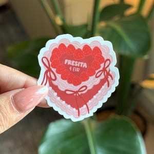 May include: A heart-shaped sticker with a strawberry cake design. The sticker is red and pink, with strawberries around the edge. The text "FRESITA 4 EVR" is printed on the sticker. A decorative sticker.
