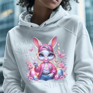 May include: White hoodie featuring a cartoon bunny wearing a pink cap, glasses, and a pink jacket. The bunny is blowing a bubble and giving a peace sign. Pink and blue Easter eggs and butterflies surround the bunny.