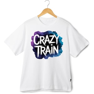 May include: White t-shirt featuring the text "CRAZY TRAIN" in white, bold lettering. The text is set against a watercolour-style background in purple and blue hues. The t-shirt is displayed on a wooden hanger.