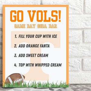 Könnte beinhalten: Ein rechteckiges Schild mit orangefarbenem Rand und dem Text "GO VOLS! GAME DAY SODA BAR." Anweisungen sind unten aufgeführt: "1. FÜLLEN SIE IHRE TASSE MIT EIS, 2. FÜGEN SIE ORANGE FANTA HINZU, 3. FÜGEN SIE SÜSSE SAHNE HINZU, 4. MIT SCHLAGSAHNE BELEGEN." Eine Football-Grafik befindet sich unten.
