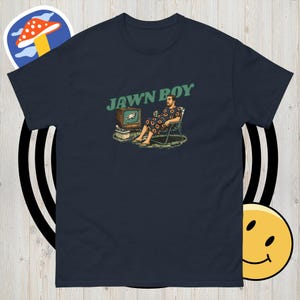 May include: Navy blue t-shirt with the text "JAWN BOY" in green. The graphic depicts a person in a floral robe sitting in a chair, watching TV. The TV shows the Philadelphia Eagles logo. A mushroom sticker is in the top left corner.