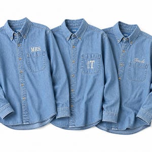 Könnte beinhalten: Drei hellblaue Jeanshemden mit Knöpfen. Auf einem Hemd ist "MRS" auf der Tasche gestickt, auf einem anderen "AT" und auf dem dritten "Bride". Jedes Hemd hat einen Button-Down-Kragen und eine einzelne Brusttasche.