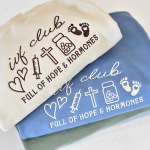 Peut inclure: Trois sweat-shirts crème, bleu et vert sont empilés. Chacun porte le texte "ivf club" et "FULL OF HOPE & HORMONES" avec des icônes graphiques de cœurs, une seringue, une croix, un flacon de pilules et des empreintes de pas de bébé.