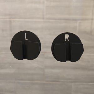May include: Two black, circular, 3D-printed objects. One has an "L" cutout, the other an "R". Each has a rectangular section below the circle. The background is a light gray, tiled surface.