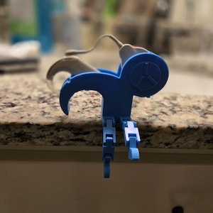 May include: A blue 3D-printed device with a circular design and a three-pronged symbol. The device has two articulated legs and a curved arm. It is resting on a speckled countertop.