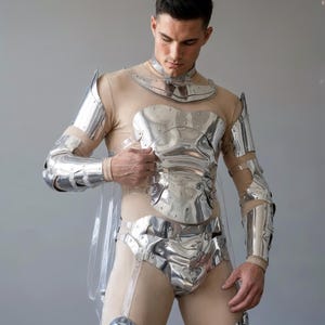 May include: A set of polished silver-toned metal armor pieces worn over a beige bodysuit. The armor includes a neck collar, a chest plate, shoulder guards, arm bracers, and a pelvic plate, all secured with clear straps and metal rivets.