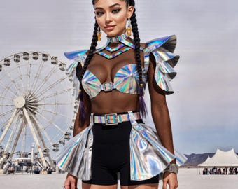 May include: A woman wearing a futuristic, iridescent silver outfit with a matching top, skirt, and shoulder pieces. The outfit is paired with black shorts and a belt. The background includes a Ferris wheel and tents, suggesting a festival setting.
