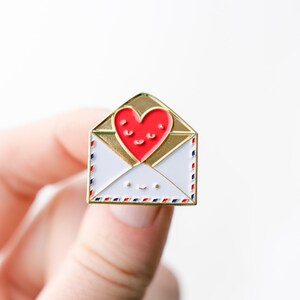 May include: Gold enamel pin shaped like an envelope with a red heart inside. The heart has a smiling face and the envelope has a smiley face.