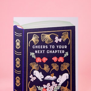 May include: A navy blue card with gold and white accents features a book with a floral and leaf design. The text on the card reads "Cheers to your next chapter."