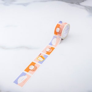 May include: A roll of washi tape with a repeating pattern of cartoon food illustrations. The tape features a white cloud, a bao bun, a slice of bread with the text "Toast!" and a stack of pancakes. The tape is on a white marble surface.
