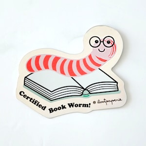 May include: A pink and red striped worm wearing glasses is on top of a closed book. The book has a teal blue spine and the text "Certified Book Worm!" is written below the book. The text "@lootpaperie" is written in a smaller font below the book.