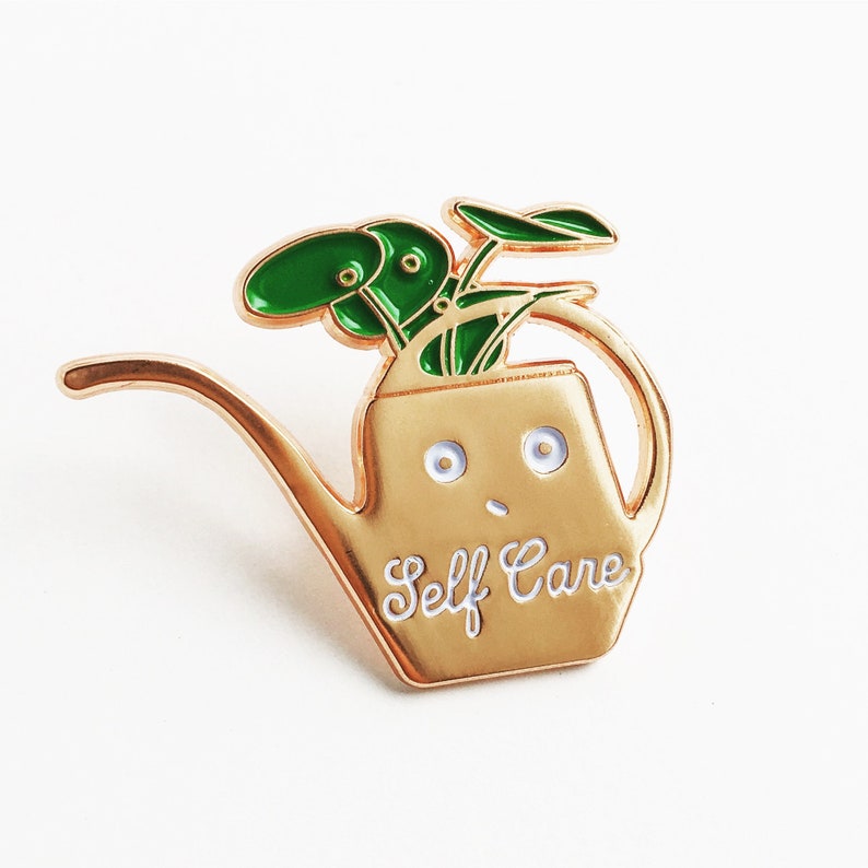 Puede incluir: Pin de esmalte dorado de un regadera con una planta verde en su interior. La regadera tiene una cara con ojos y boca y el texto "Self Care" en la parte delantera.