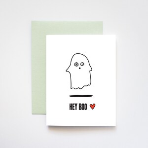 May include: A white card with a black and white illustration of a ghost with a surprised expression. The text "HEY BOO" is written in black below the ghost with a red heart.