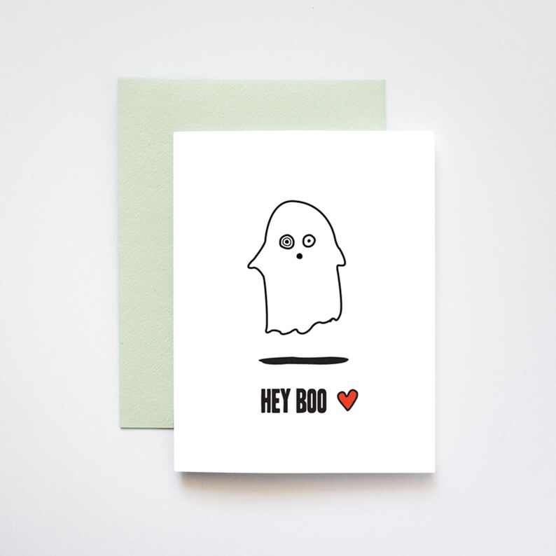 May include: A white card with a black and white illustration of a ghost with a surprised expression. The text "HEY BOO" is written in black below the ghost with a red heart.