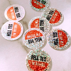 May include: A collection of eight round, holographic stickers with the text "Hell Yes I'm Voting" in black and red. The stickers have a glittery, iridescent finish.